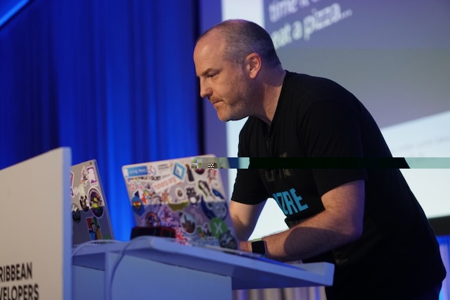 A picture of Jim on a stage at a conference standing next to a podium with a laptop on it. Jim is wearing a black t-shirt and is working on the laptop